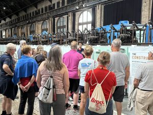  A group of 14 people listen to a tour guide speak while standing in front of large pieces of equipment that were used in the past to generate power. 