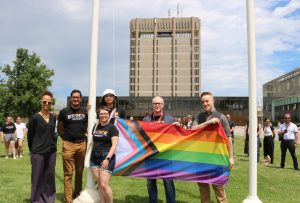 A group of people holds a colourful Pride flag beside flagpoles in front of a concrete tower.