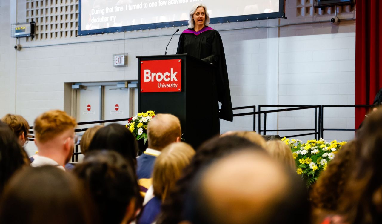 A woman in academic regalia speaks at a podium during a university graduation ceremony.