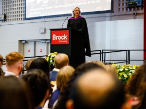 A woman in academic regalia speaks at a podium during a university graduation ceremony.