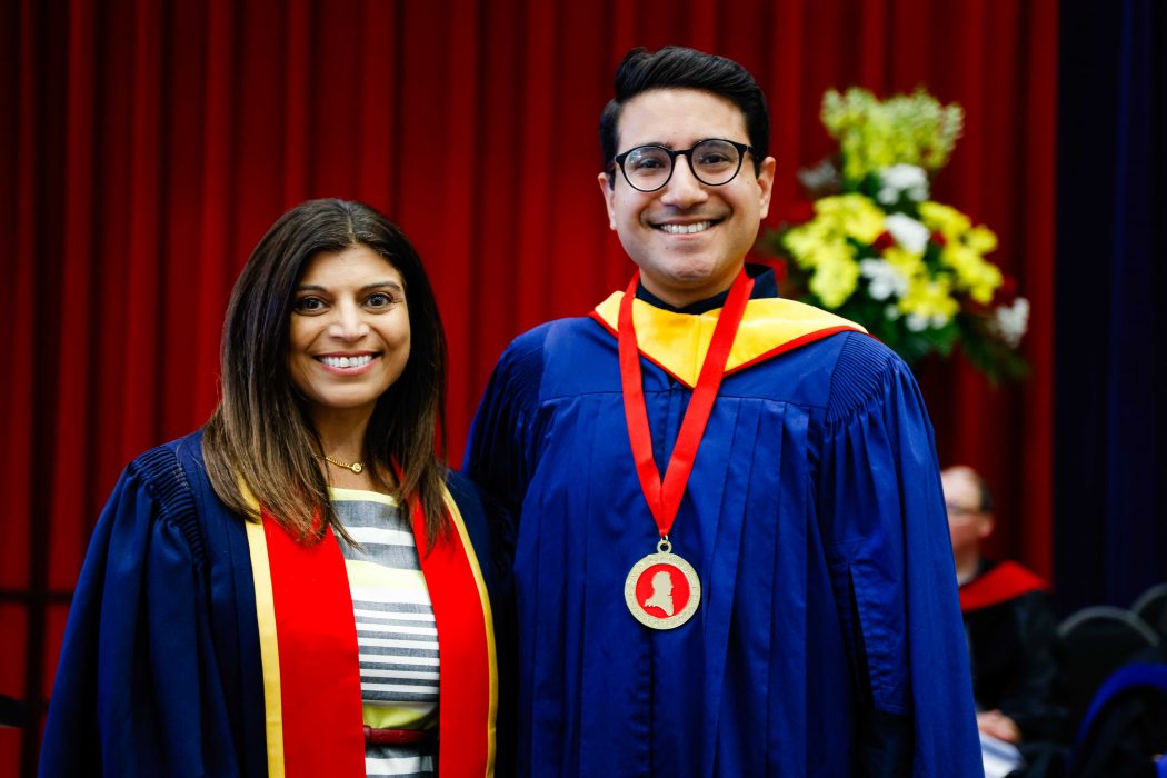 A young man wearing a blue academic robe and a medal poses for a photo next to a woman in academic regalia.