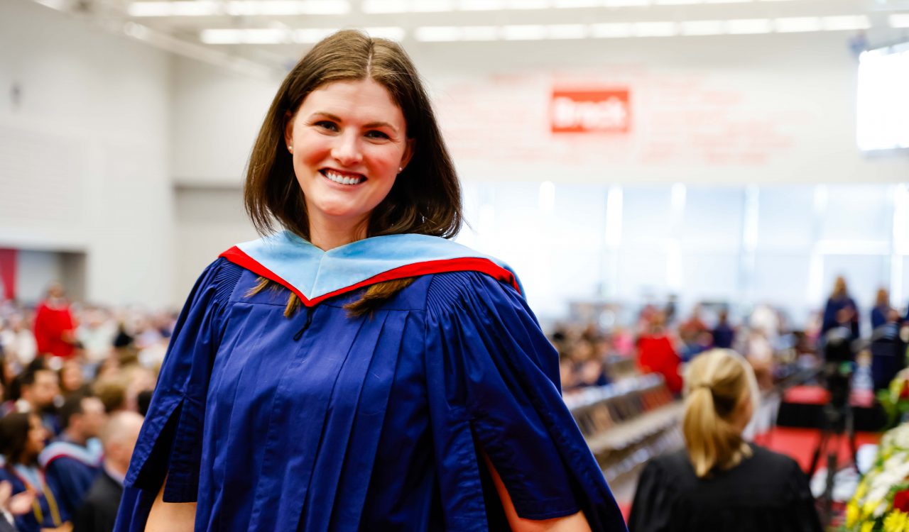 A woman wearing a graduation gown stands in front of a gymnasium filled with graduates.