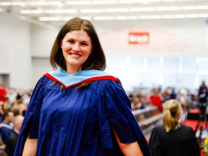 A woman wearing a graduation gown stands in front of a gymnasium filled with graduates.