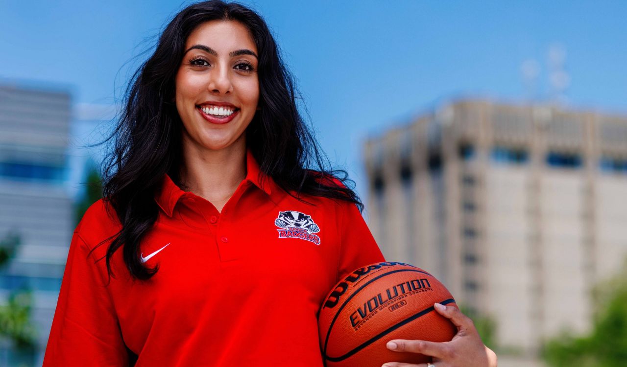 A woman stands outdoors at Brock University’s campus while smiling and wearing a bright red polo while holding a basketball under her arm.