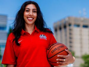 A woman stands outdoors at Brock University’s campus while smiling and wearing a bright red polo while holding a basketball under her arm.