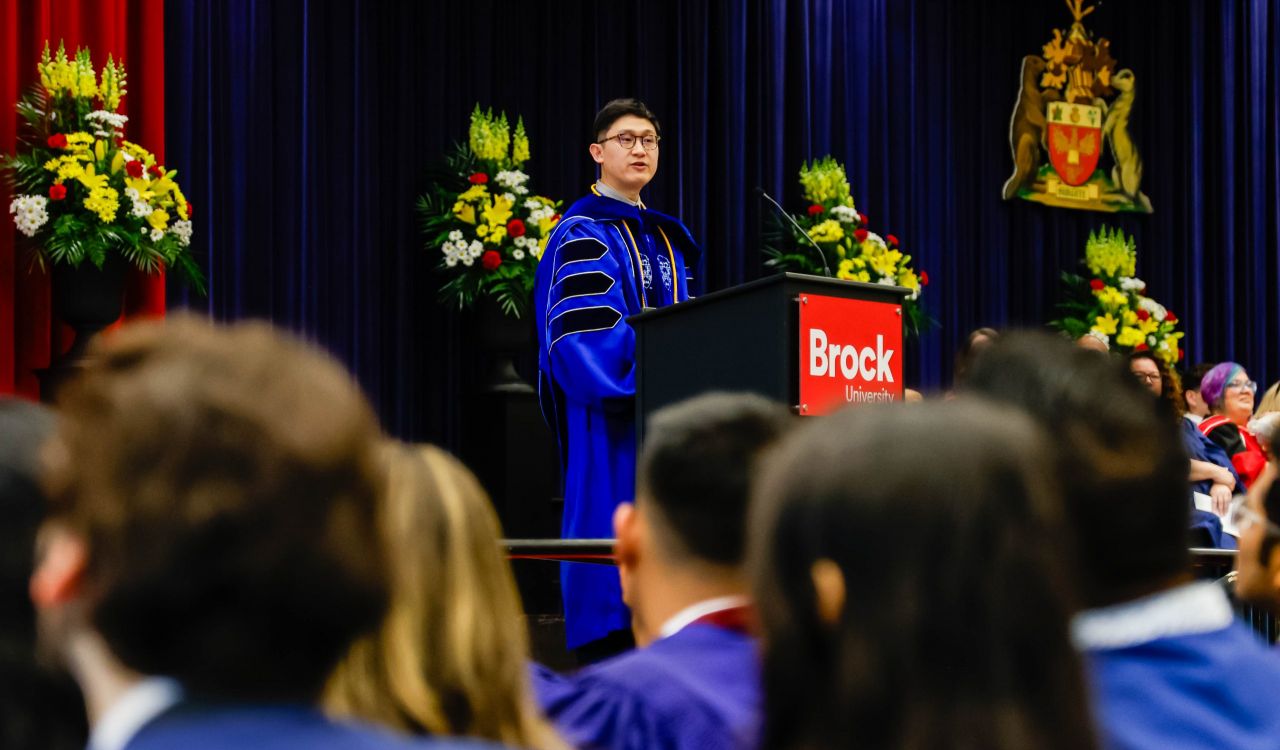 A man in academic regalia addresses an audience from a podium.