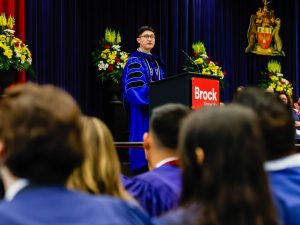 A man in academic regalia addresses an audience from a podium.