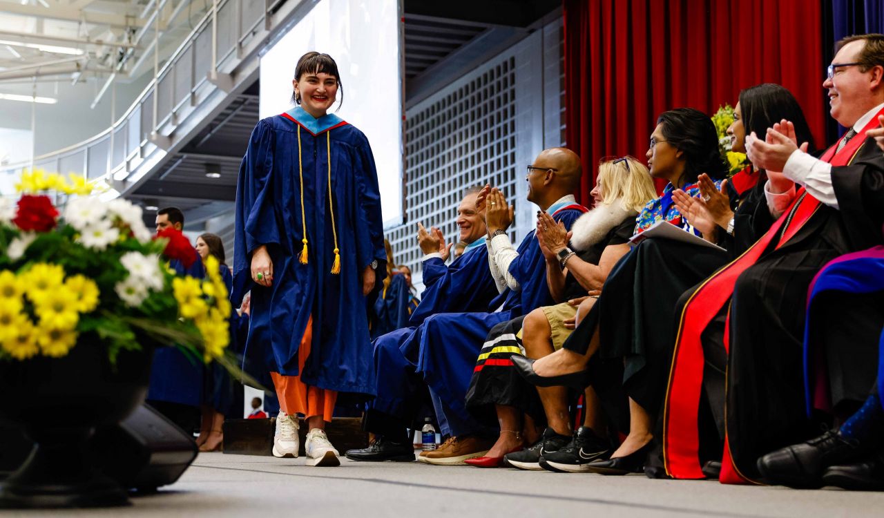 A woman wearing a graduation gown walks across a Convocation stage as the seated platform party looks on.