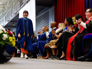 A woman wearing a graduation gown walks across a Convocation stage as the seated platform party looks on.