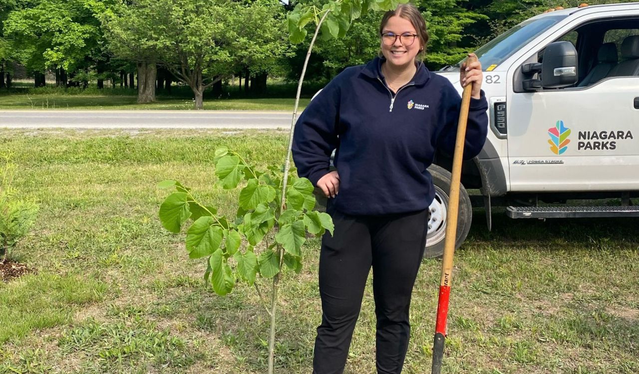 Sydney McIntyre stands outside with a shovel in her hand behind a newly planted tree.