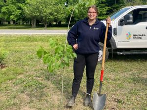 Sydney McIntyre stands outside with a shovel in her hand behind a newly planted tree.