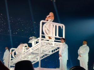 A woman sings on stage in a white dress while standing on a white platform.