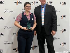 Two people pose for a photo in front of a logo backdrop. One of them is holding an award.