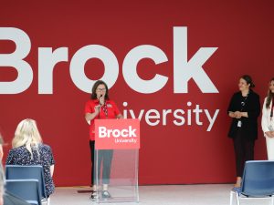 A woman stands in front of an audience speaking while behind a podium. Two women standing to the side look on. 