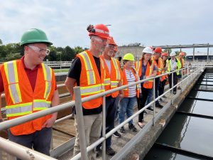 A group of people in orange high-visibility vests and hard hats look over an outdoor railing that leads to water.