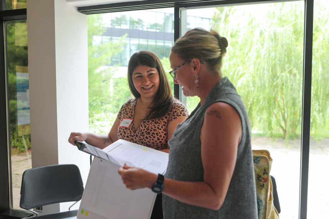 Two women stand looking at data in binders in front of a window. 