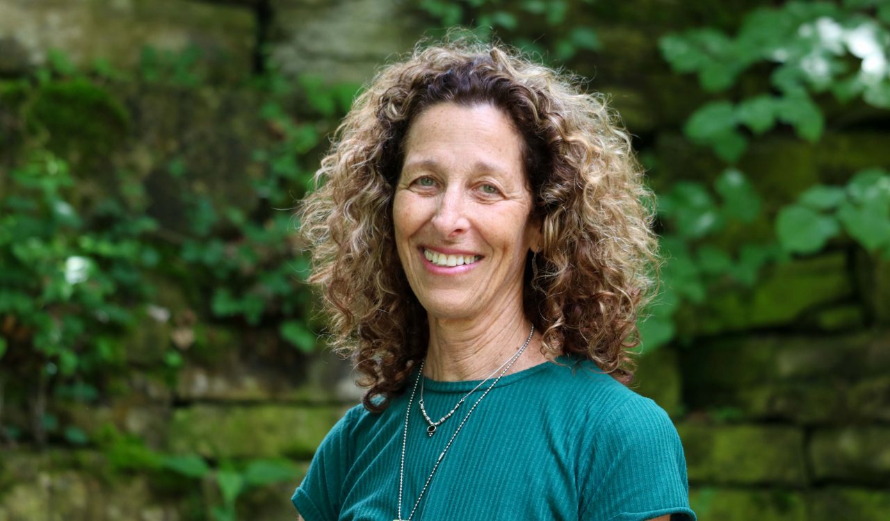 A woman poses for a portrait outside with greenery and a stone wall behind her.