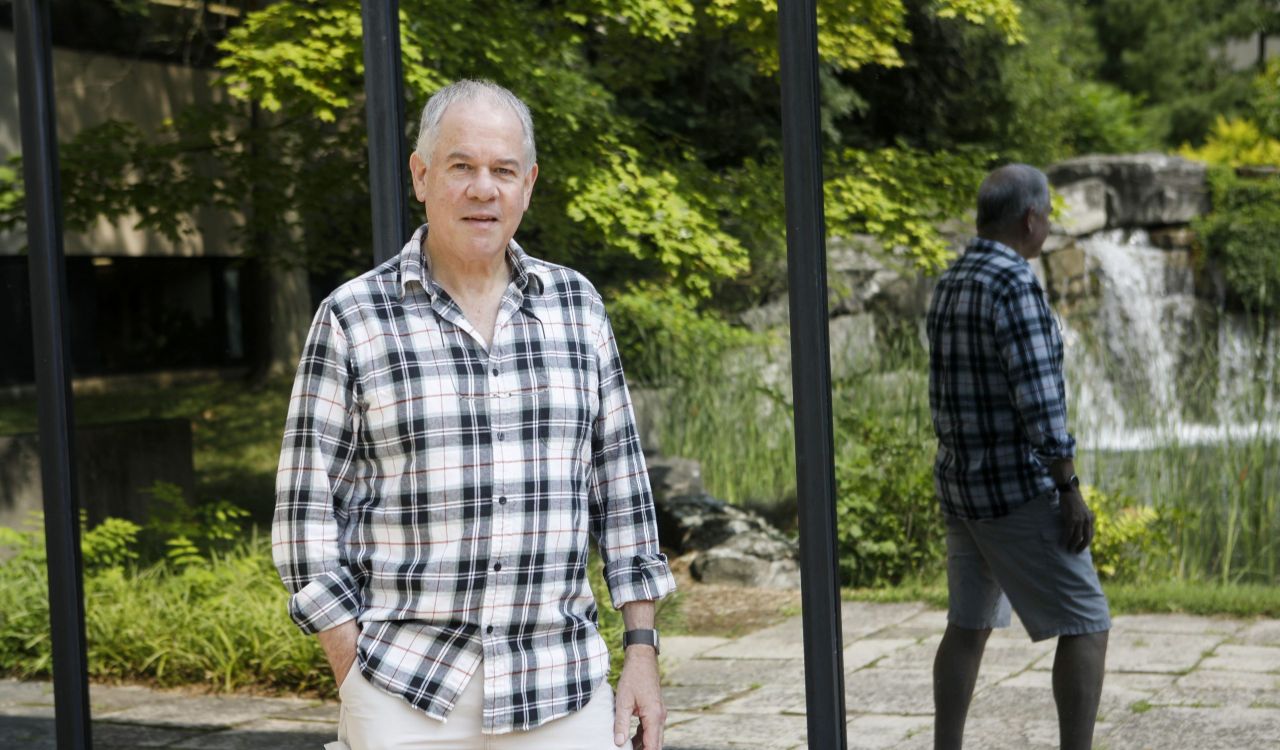 Professor Martin Head stands outside the Pond Inlet at Brock University. His back is to mirrored glass, so you can see his reflection as well as the reflection of a pond and waterfall.