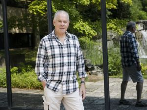 Professor Martin Head stands outside the Pond Inlet at Brock University. His back is to mirrored glass, so you can see his reflection as well as the reflection of a pond and waterfall.