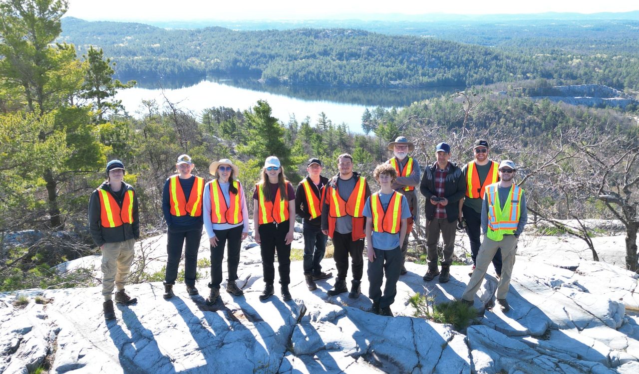 Small group of Brock University Earth Sciences students and professor stand in a line on a large rock formation in front of a lake.