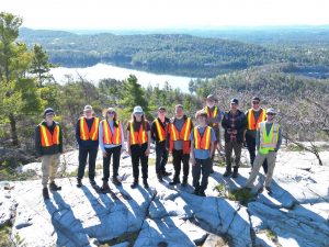 Small group of Brock University Earth Sciences students and professor stand in a line on a large rock formation in front of a lake.