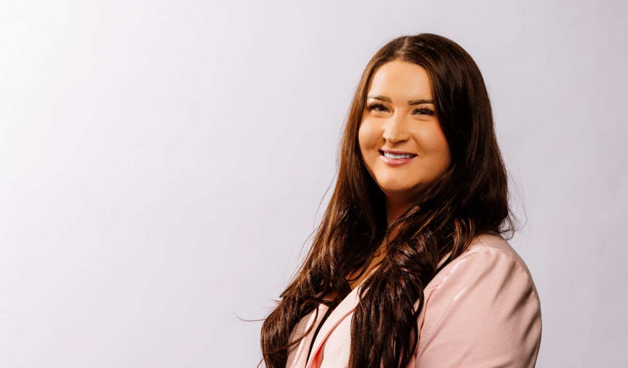 A young woman in a pink blazer poses for a portrait against a light backdrop.