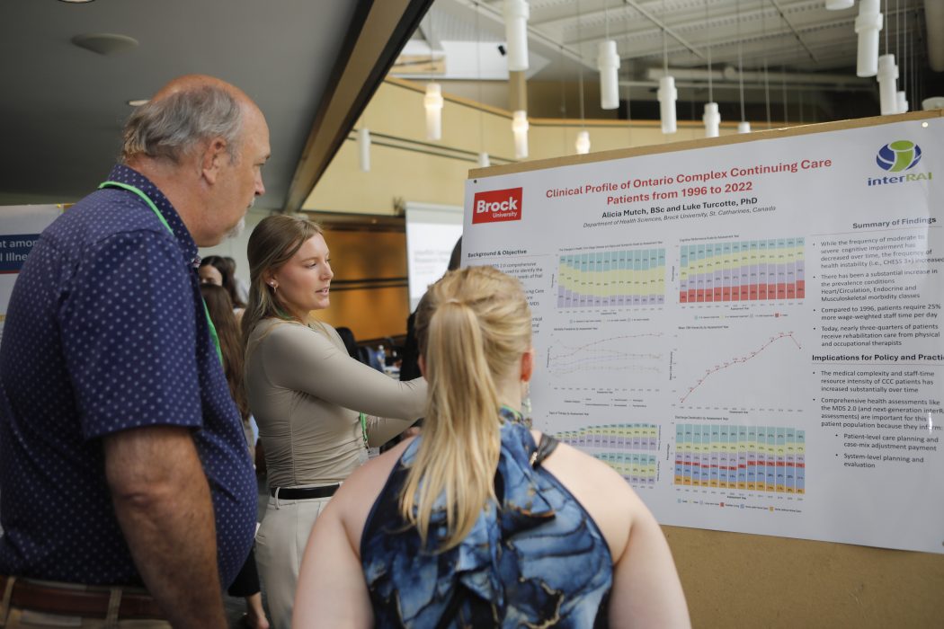 Brock University student Alicia Mutch points to a poster displaying her research, titled “Clinical Profile of Ontario Complex Continuing Care Patients from 1996 to 2022.” Two people stand next to her as they listen to her speak and look at her poster.