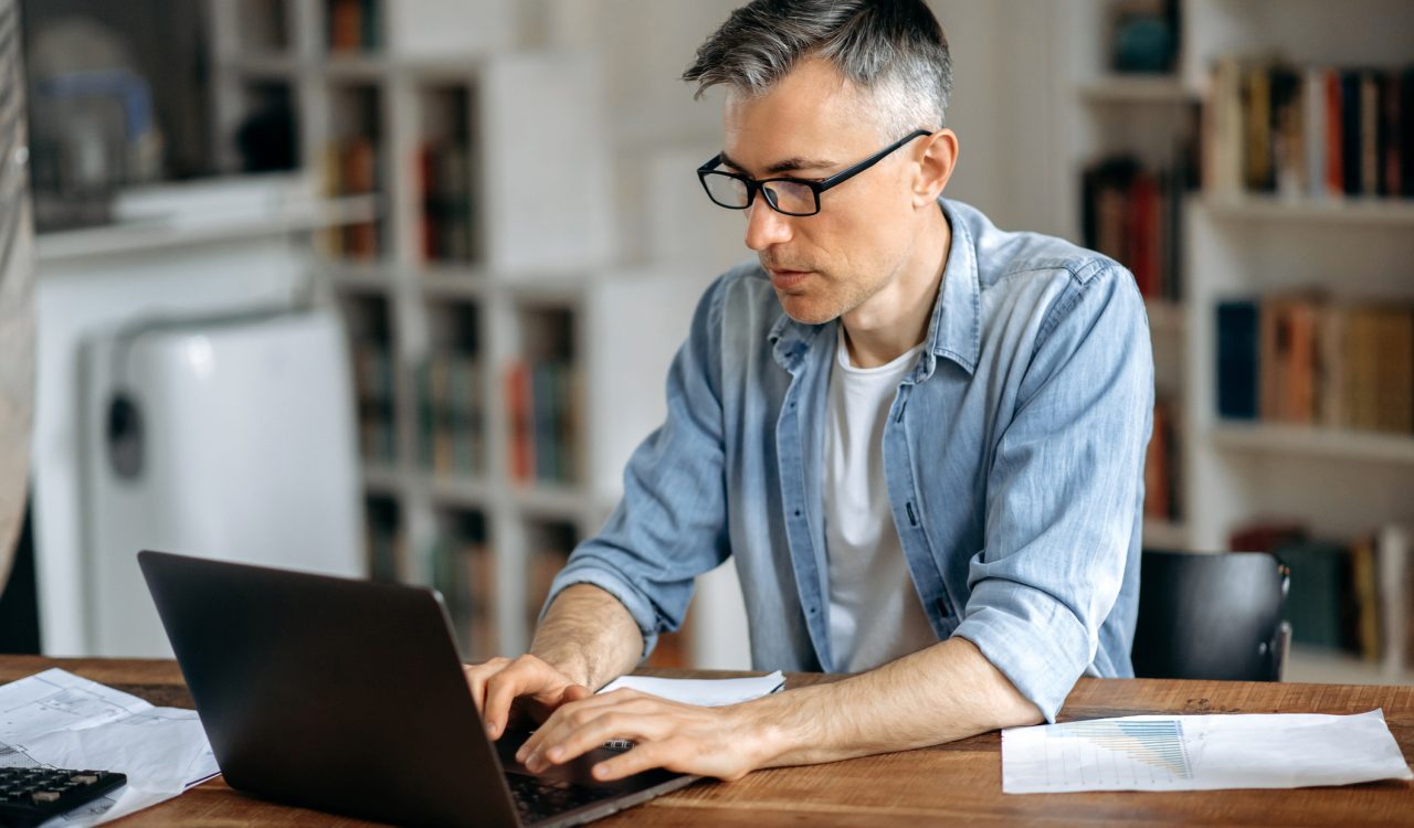 An older adult man works on a laptop in a living room.