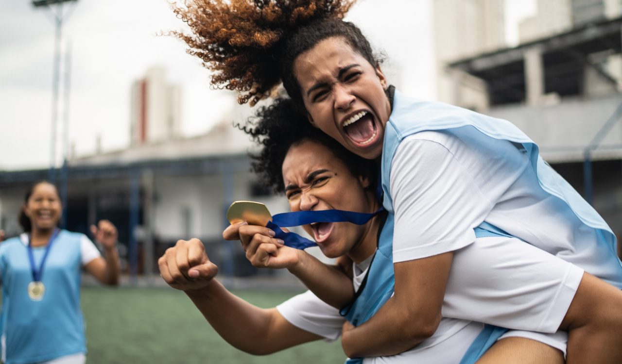 Female soccer players hold up medals and hug on a field.