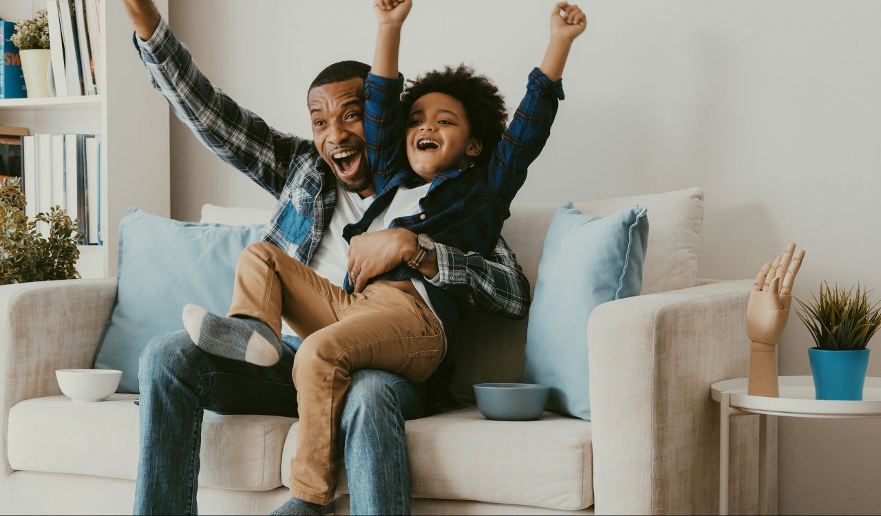 A father and son cheer while sitting on a sofa.