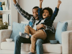 A father and son cheer while sitting on a sofa.