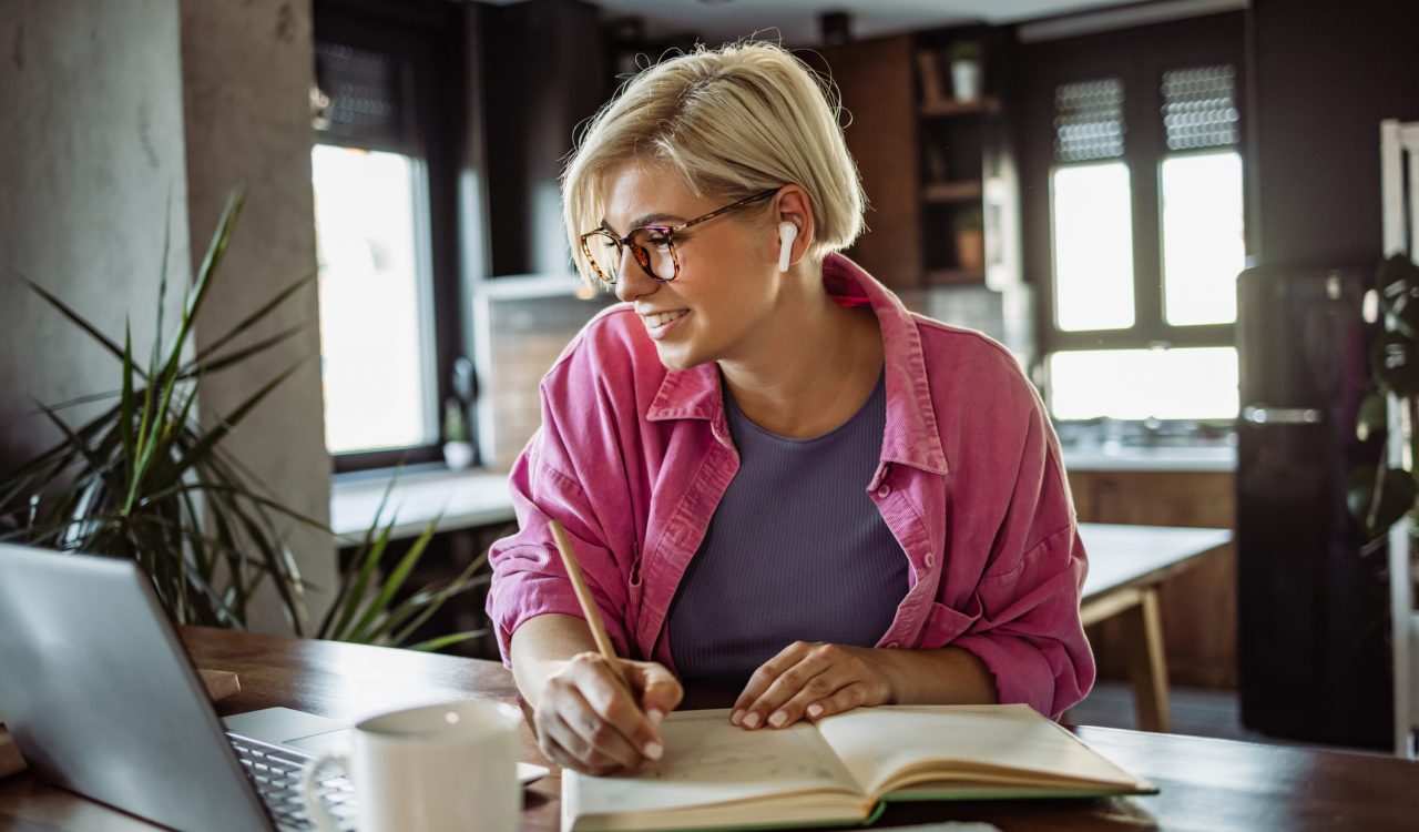 A smiling young woman in glasses takes notes in a notebook while looking at a laptop screen. She is sitting at a table in a kitchen.