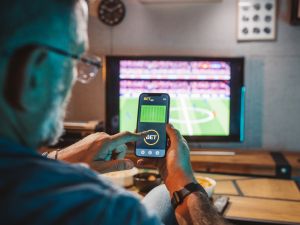 Close-up photo over the shoulder of an older man who is looking at a sport betting app on his phone with a sporting event on the television in the background.