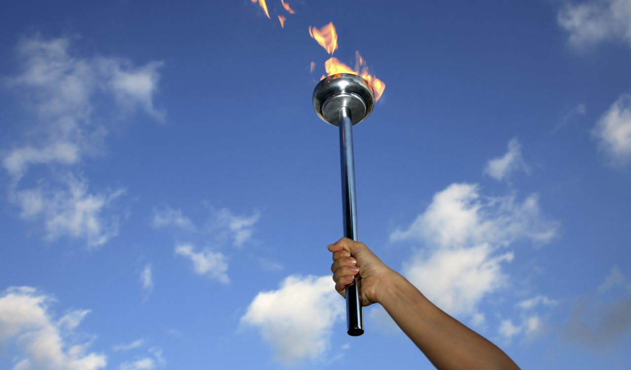 A close-up of a hand holding up a silver Olympic flame torch against a blue sky.