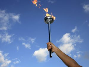 A close-up of a hand holding up a silver Olympic flame torch against a blue sky.