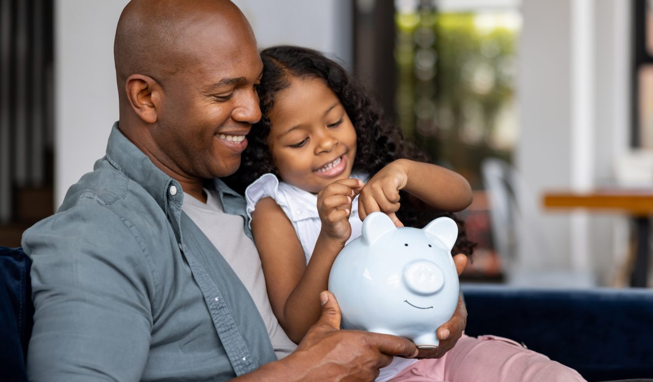 A father sits with his daughter on a sofa. She is smiling and putting money in a piggy bank.