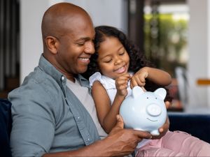 A father sits with his daughter on a sofa. She is smiling and putting money in a piggy bank.