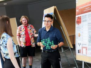 Brock University Physics student Sebastien Duguay holds a 3D printed crystal structure as they speak to a fellow student about their research on “3D Printing Crystal Structures for Education and Research.” Behind them is a large poster outlining their research.