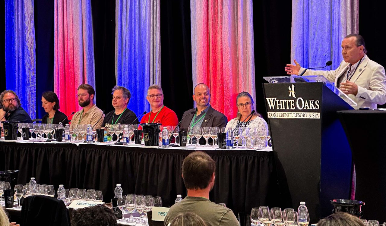 A group of people sit at a long table in front of an audience while a man speaks from a podium during a conference event.