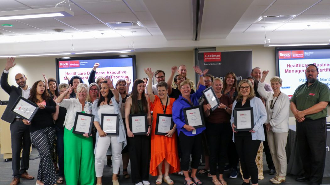 A group of people holding framed certificates pose for a photo in a meeting room.