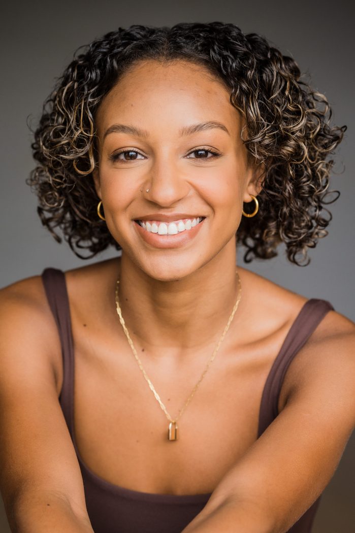 A portrait headshot shoes a young Black woman with curly hair from the shoulders up. She is smiling warmly at the camera with gold hoop earrings and a gold necklace.