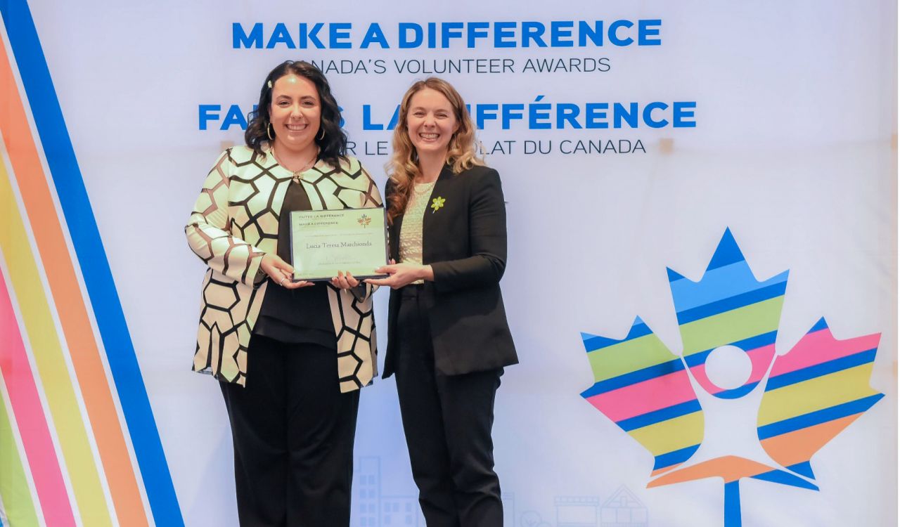 Lucia Marchionda stands beside the Jenna Sudds, Member of Parliament for Kanata — Carleton, in front of a banner with a multicoloured maple leaf. Both women are wearing business clothes and smiling while holding a certificate.