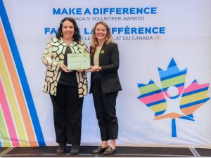 Lucia Marchionda stands beside the Jenna Sudds, Member of Parliament for Kanata — Carleton, in front of a banner with a multicoloured maple leaf. Both women are wearing business clothes and smiling while holding a certificate.