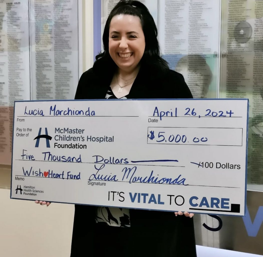 Lucia Marchionda stands at McMaster Children’s Hospital holding an over-sized donation cheque in the amount of $5,000. She has black hair and is smiling warmly.