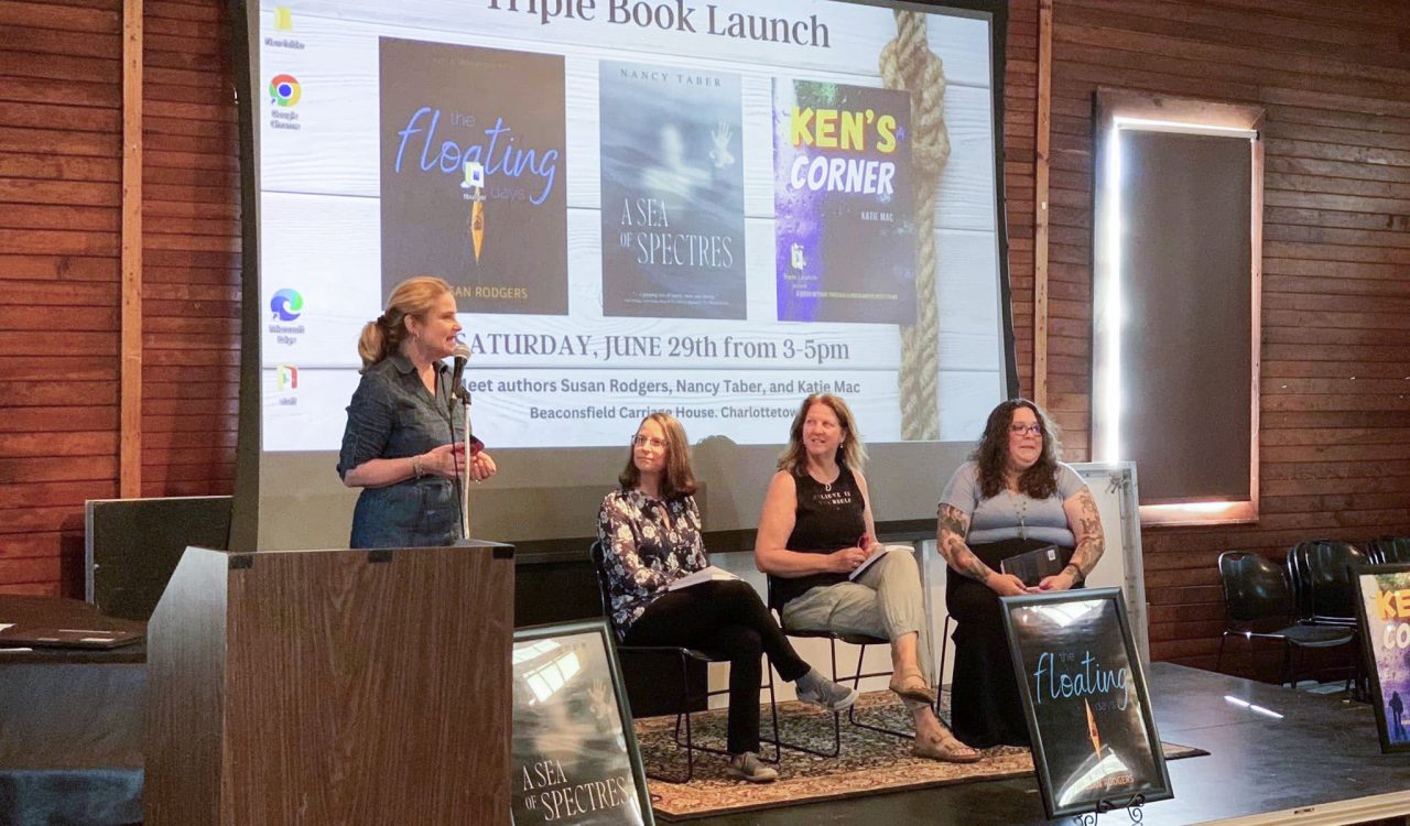 A group of women sit on a stage in front of a large screen showing three book covers. One woman stands in front of a microphone while the other women look on.