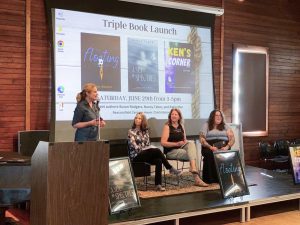 A group of women sit on a stage in front of a large screen showing three book covers. One woman stands in front of a microphone while the other women look on.