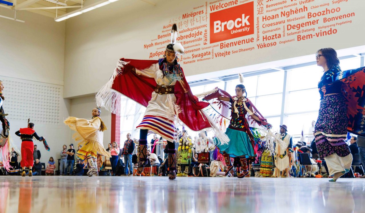 A group of women dance in traditional Indigenous regalia at a pow wow in a sunny gymnasium.