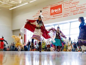 A group of women dance in traditional Indigenous regalia at a pow wow in a sunny gymnasium.