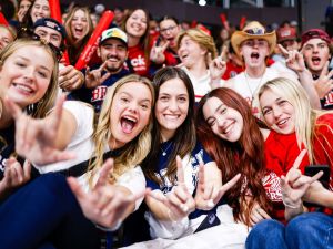 A crowd of people with excited expressions on their faces raise their hands in celebration while sitting in a hockey arena.