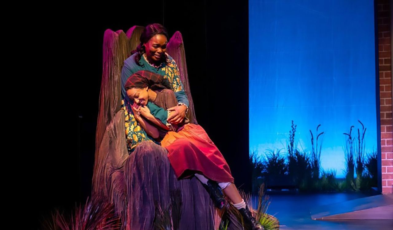 A Black woman cradles a younger Black woman in her arms on stage. The two women are wearing 1850’s period costumes and sit on a chair that looks like a tree. The lighting is low and moody. The younger person smiles, the older person appears to be crying.